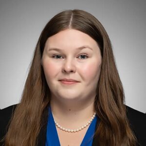 A professional headshot of Sidney Lewellen, featuring her with long, straight brown hair, wearing a blue blouse, a black blazer, and a pearl necklace, set against a neutral background.